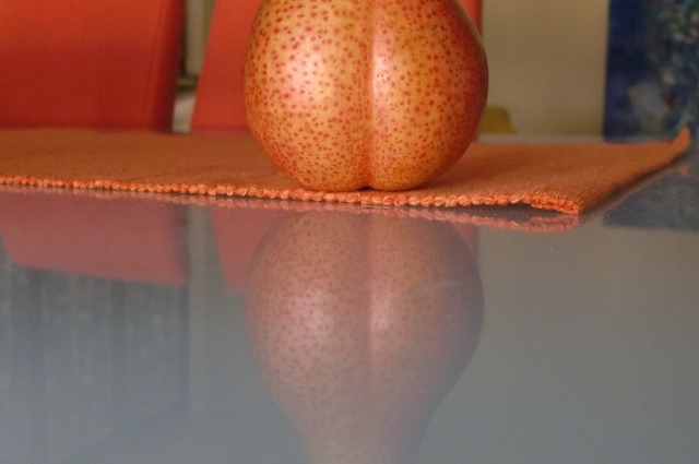 Auf dem Foto sieht man einen Teil einer Forellenbirne, die sich im Glastisch spiegelt. Ihre Rundungen erinnern an einen sexy Po. Die Forellenbirne ist gelbgrün, mit einer rötlichen Seite und hat wunderschöne orange-rote Sprenkel.